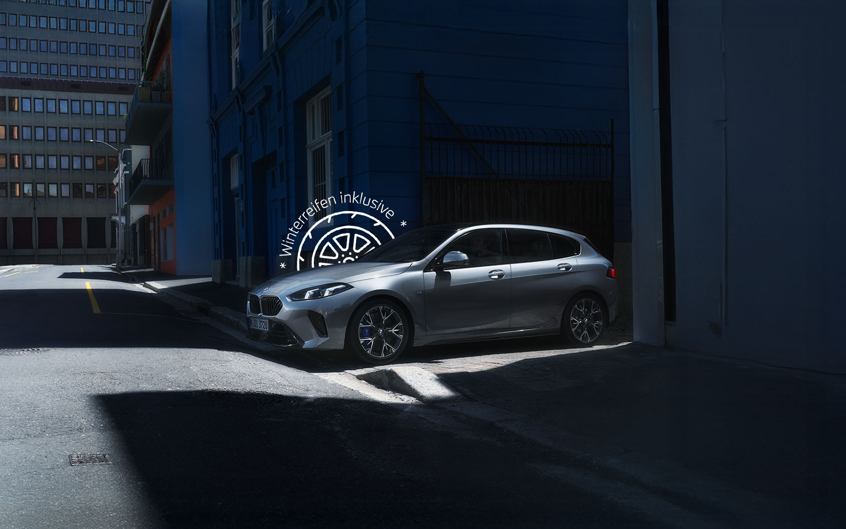 Silberner BMW 1er parkt im Schatten einer städtischen Hausfassade, Werbehinweis "Winterreifen inklusive" über dem Fahrzeug