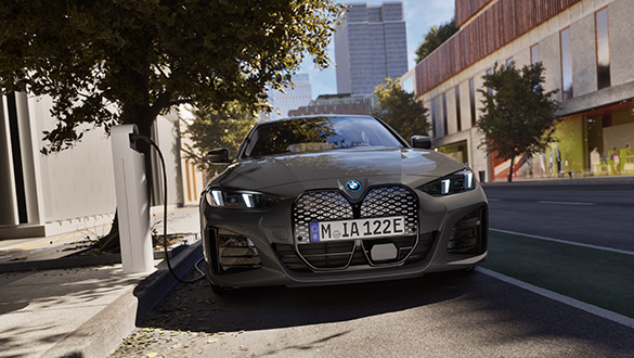 Frontansicht eines dunkelgrauen BMW i4 im städtischen Raum angeschlossen an einer Ladesäule neben einem Baum an einer Straße, Fokus auf Grill & Scheinwerfer