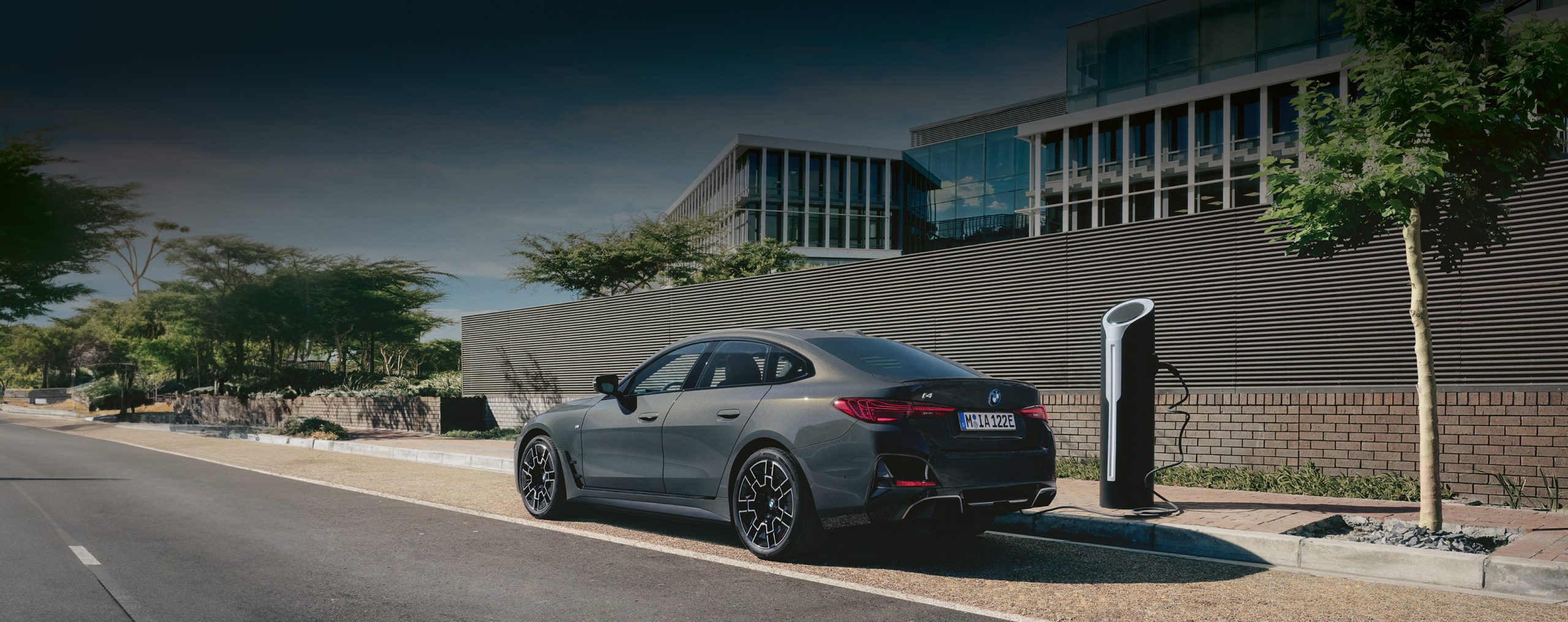 Grauer BMW i4 in Seitenansicht angeschlossen an einer Ladesäule neben einer Straße vor einem verglasten Bürogebäude