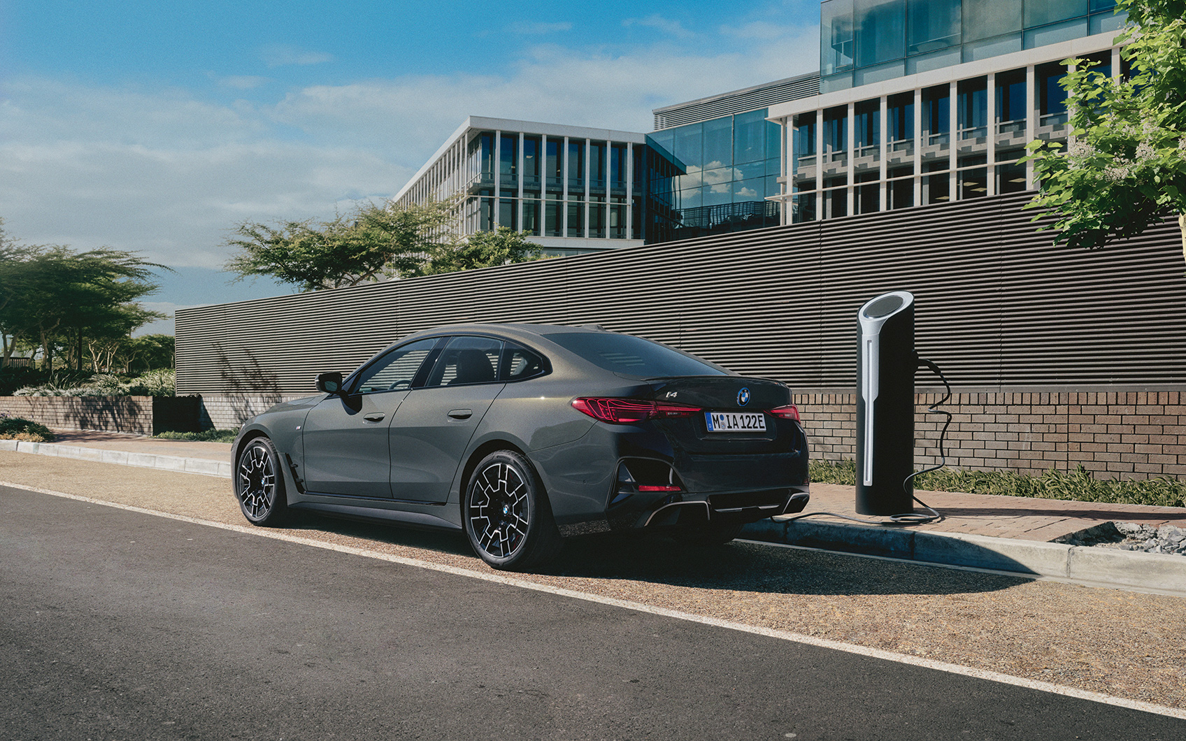 Grauer BMW i4 in Seitenansicht angeschlossen an einer Ladesäule neben einer Straße vor einem verglasten Bürogebäude