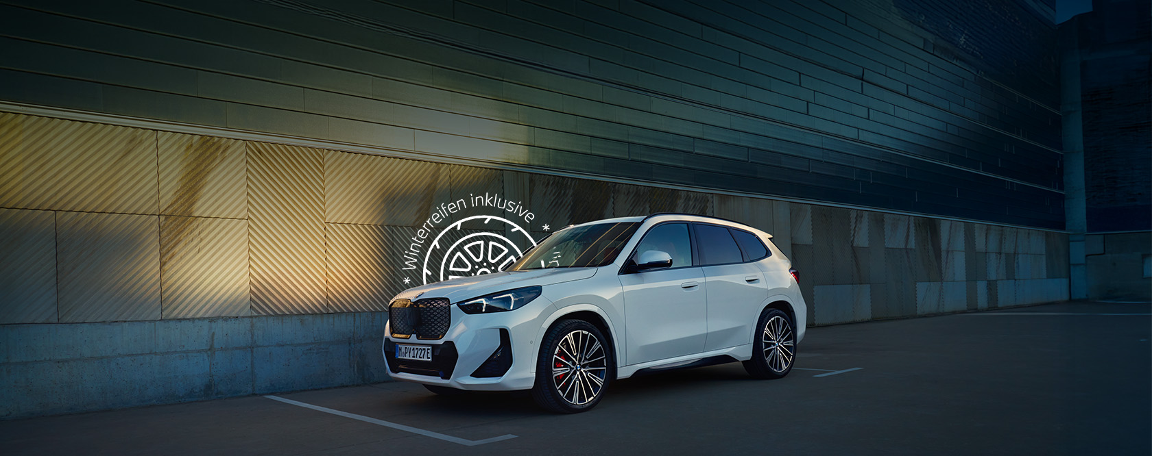 Weißer BMW iX1 steht auf einem Parkplatz vor einer modernen Metallfassade, Werbehinweis „Winterreifen inklusive“ über dem Fahrzeug.