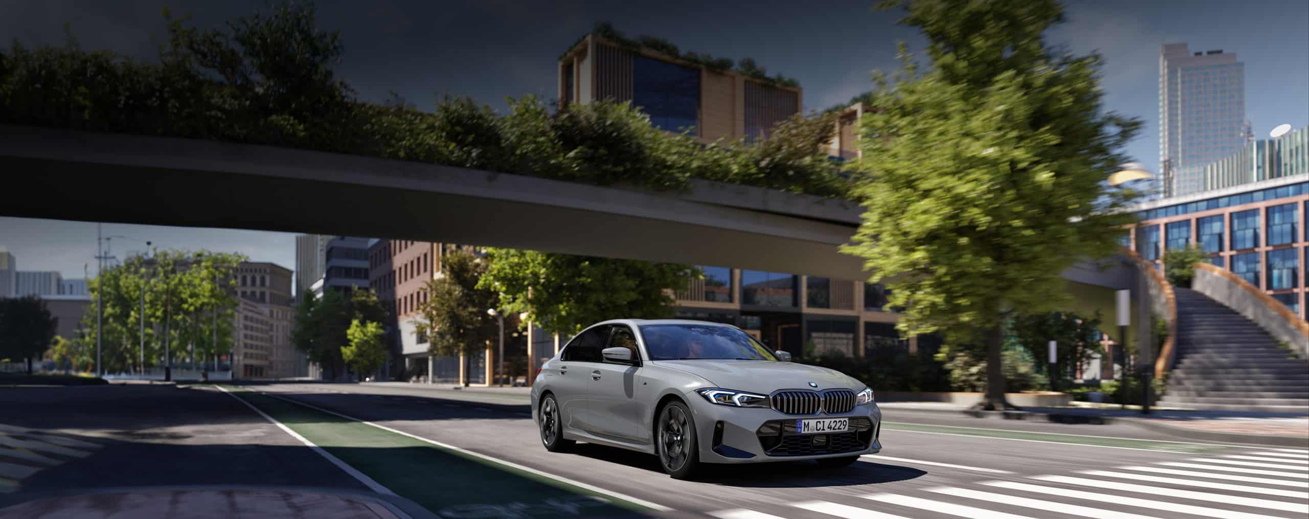 Graue BMW 3er Limousine fährt über eine städtische Straße auf den Betrachter zu, Blick durch eine Fußgängerbrücke auf städtische Gebäude im Hintergrund