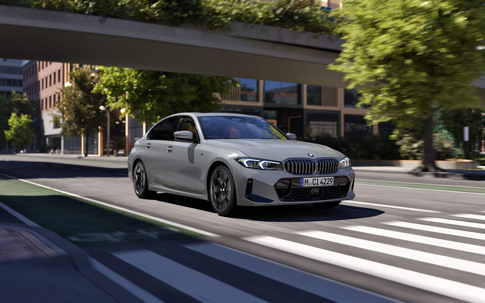 Graue BMW 3er Limousine fährt über eine städtische Straße auf den Betrachter zu, Blick durch eine Fußgängerbrücke auf städtische Gebäude im Hintergrund