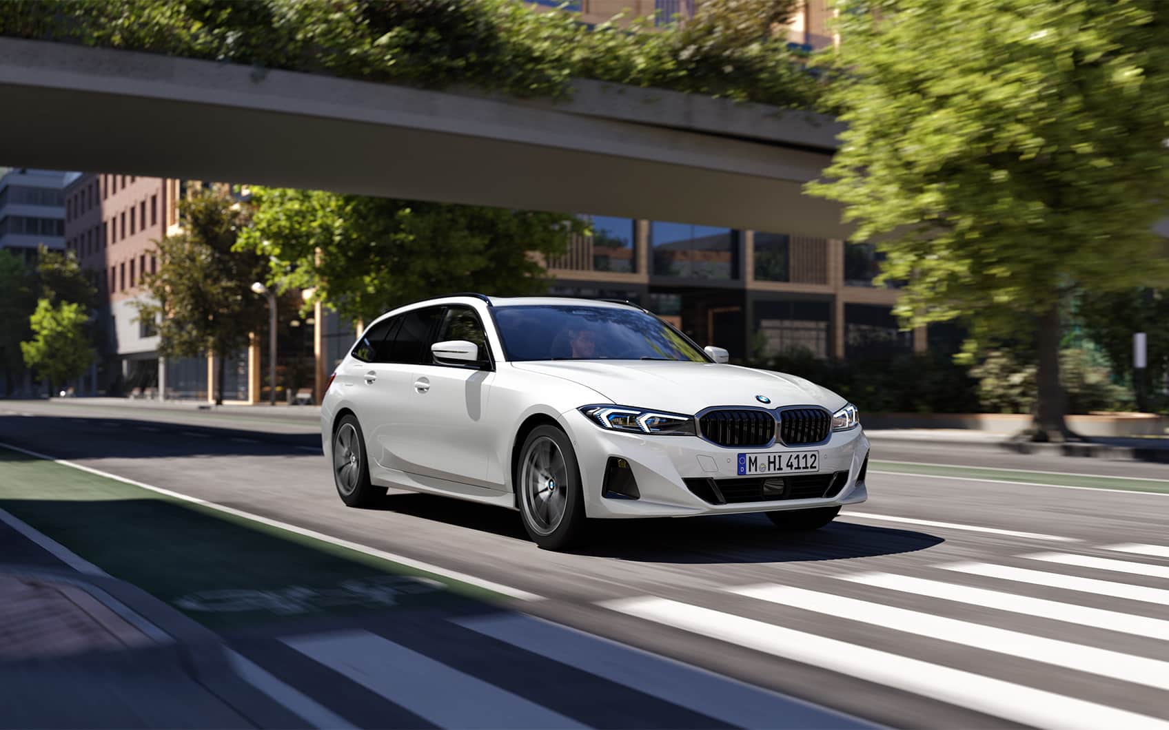 Weißer BMW 3er Touring fährt über eine städtische Straße auf den Betrachter zu, Blick durch eine Fußgängerbrücke auf städtische Gebäude im Hintergrund