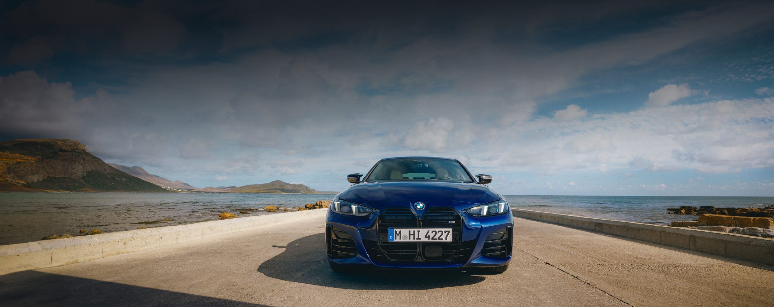 Frontaufnahme eines BMW 4er Gran Coupé M in der Farbe dunkelblau auf einer Strasse mit Meer im Hintergrund, im Fokus sind die Scheinwerfer und die BMW Niere