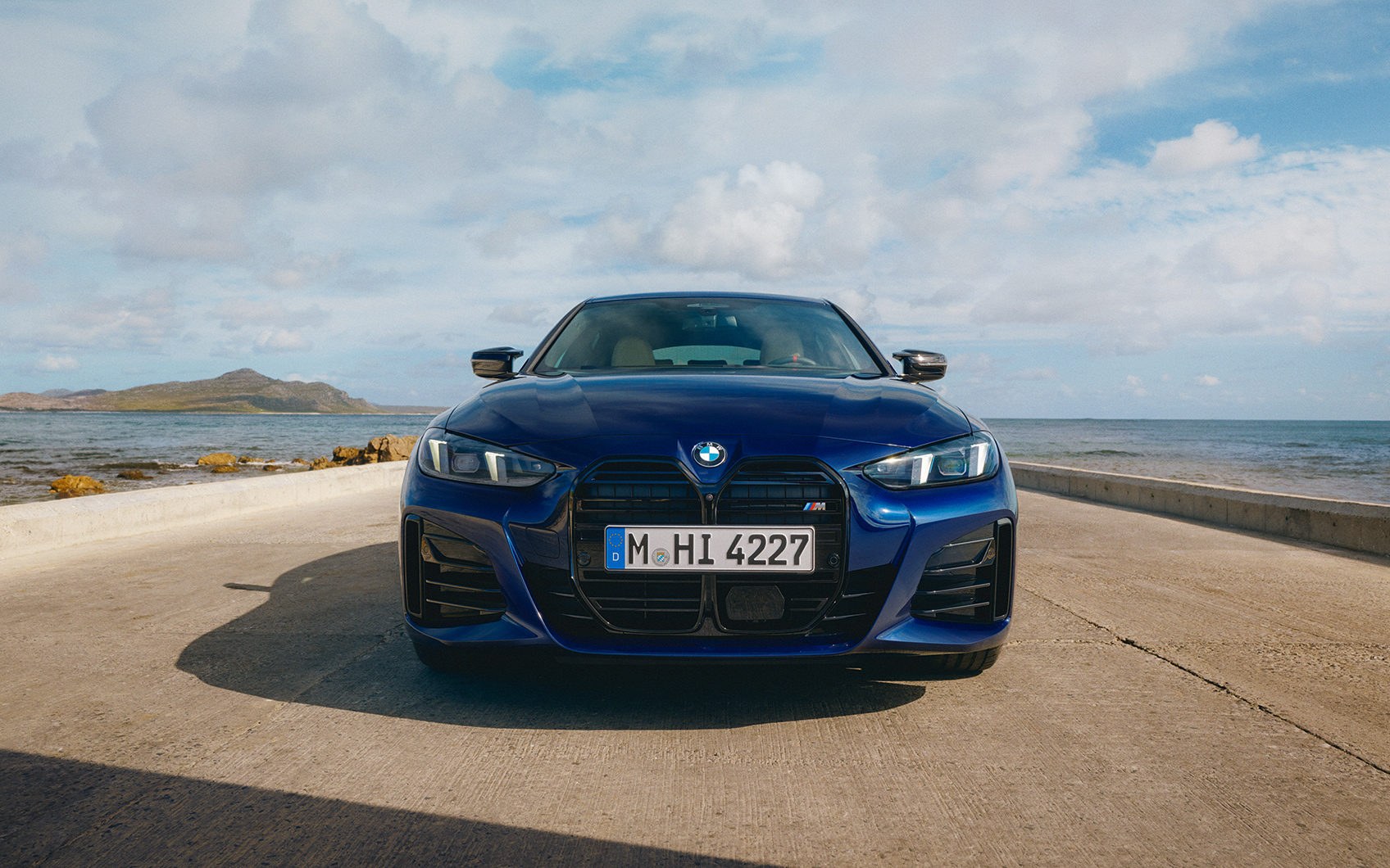 Frontaufnahme eines BMW 4er Gran Coupé M in der Farbe dunkelblau auf einer Strasse mit Meer im Hintergrund, im Fokus sind die Scheinwerfer und die BMW Niere