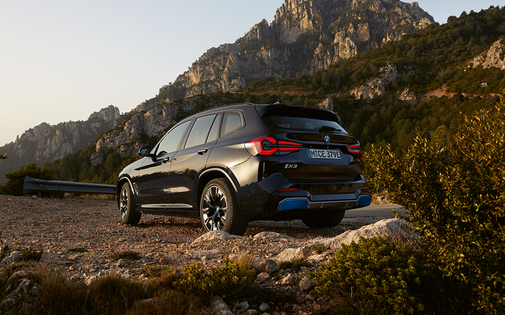 Vollelektrischer BMW iX3 SUV in bergiger Landschaft, seitlich von hinten beleuchtet