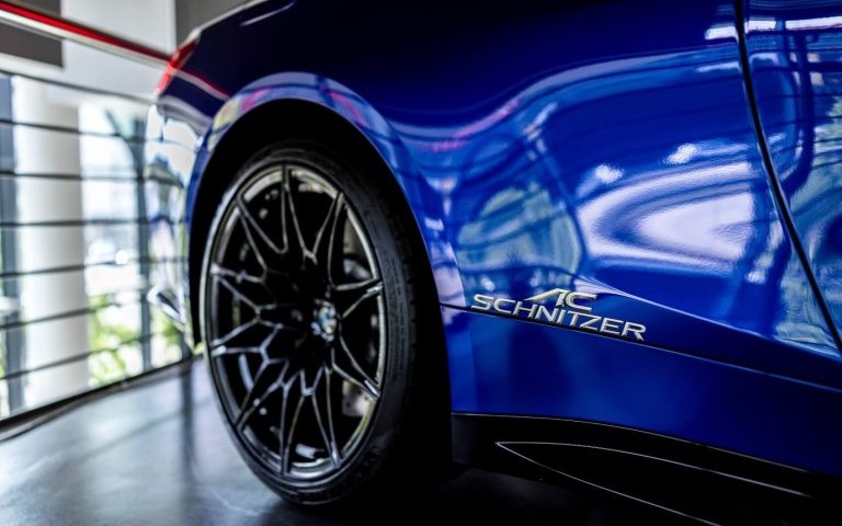 Detailansicht des rechten Hinterreifens eines BMW M4 Competition Coupé in der Farbe Dunkelblau mit Fokus auf den AC Schnitzer Schriftzug neben dem Reifen, aufgenommen im Showroom der Automag