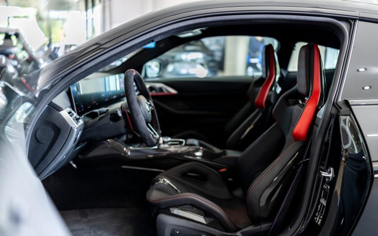 Interieur im vorderen Bereich eines BMW M4 Coupé mit Fokus auf die BMW M Sportsitze des Fahrers und des Beifahrers und den Farben Rot und Schwarz, aufgenommen durch die geöffnete Fahrertür im Showroom der Automag