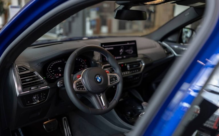 Cockpit des BMW X4 M Competition mit Fokus auf dem Lederlenkrad und den Bedienelementen, aufgenommen durch die geöffnete Fahrertür im Showroom der Automag