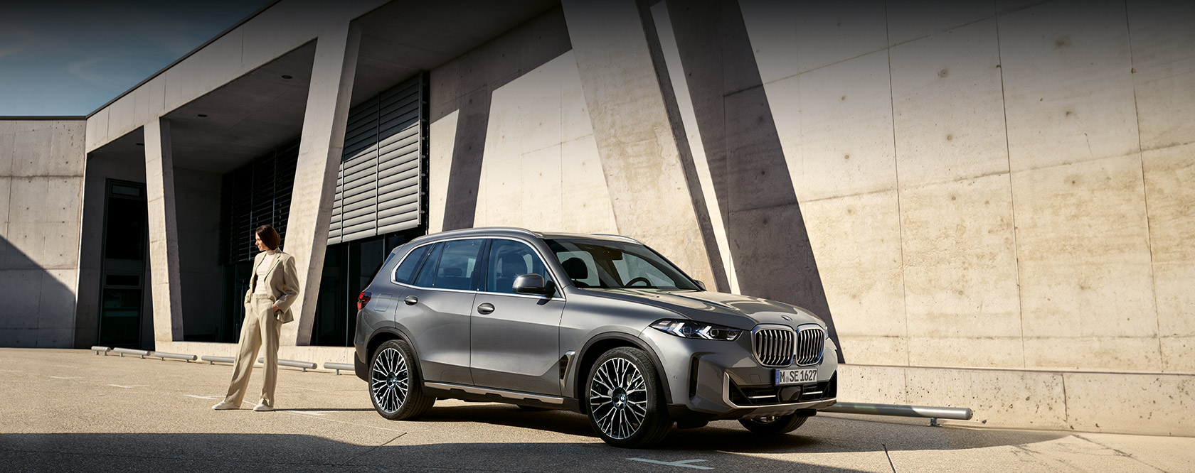 Seitenansicht des BMW X5 in der Farbe Dunkelgrau vor einem modernen Gebäude, neben dem Fahrzeug steht eine modern gekleidete Frau