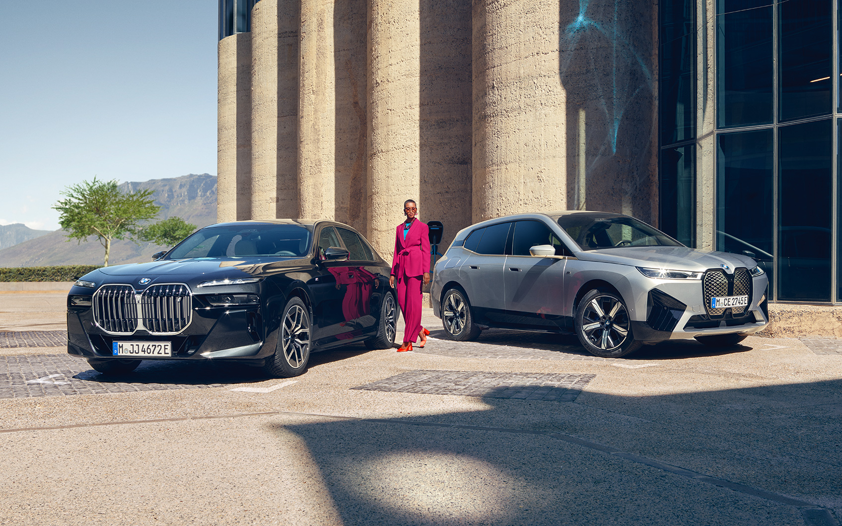 BMW 7er Limousine PHEV in der Farbe Schwarz und X7 PHEV in der Farbe Grau geparkt vor einem Gebäude; zwischen den beiden Modellen steht eine Frau in einem pinken Anzug