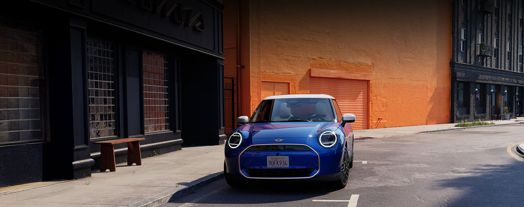 Blauer Mini Cooper Electric mit weißem Dach frontal geparkt vor orangefarbener Wand.