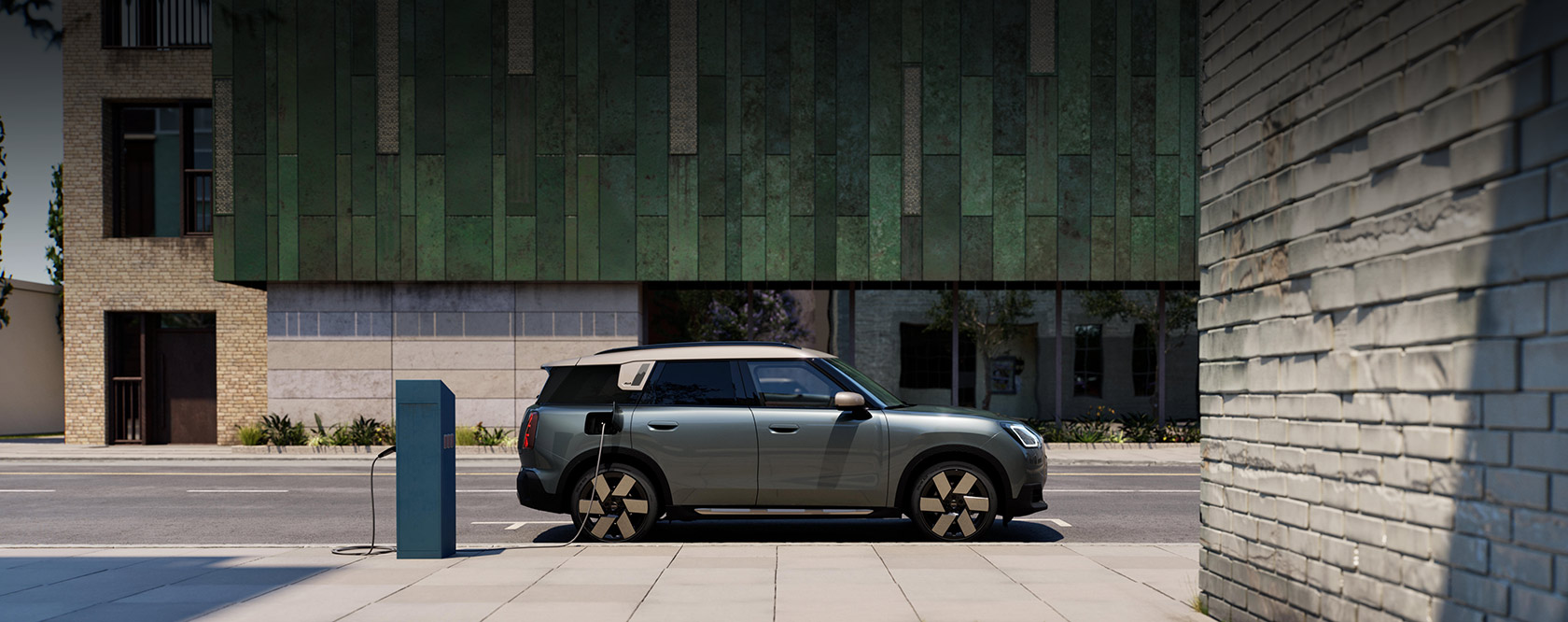 Grüner Mini Countryman Electric mit weißem Dach an Ladesäule auf städtischem Gehweg.