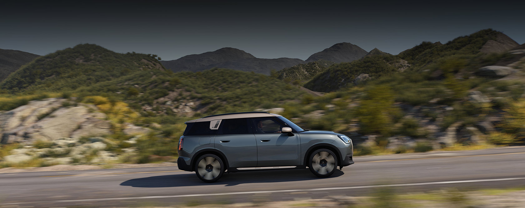 Grüner Mini Countryman mit weißem Dach fährt auf Landstraße vor Bergkulisse.