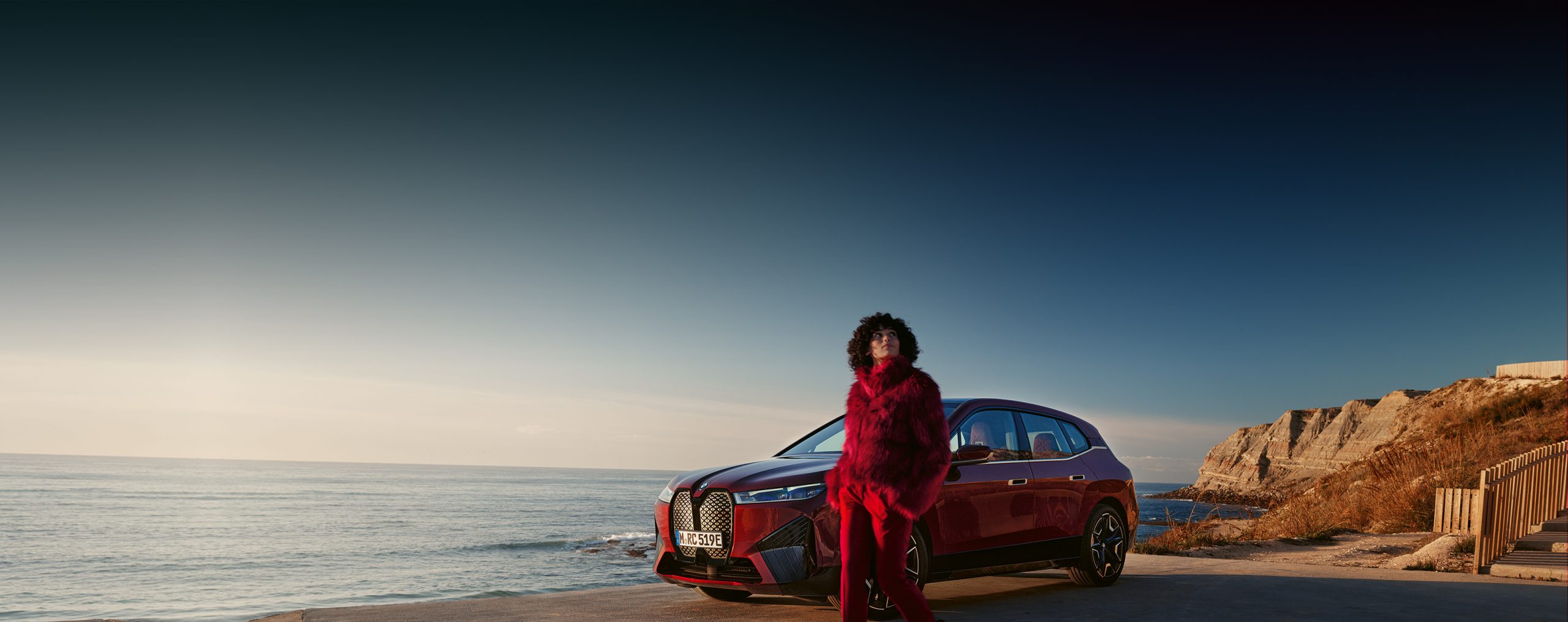 Rotes BMW iX Modell an Küstenstraße, Person in rotem Outfit, Meer und blauer Himmel