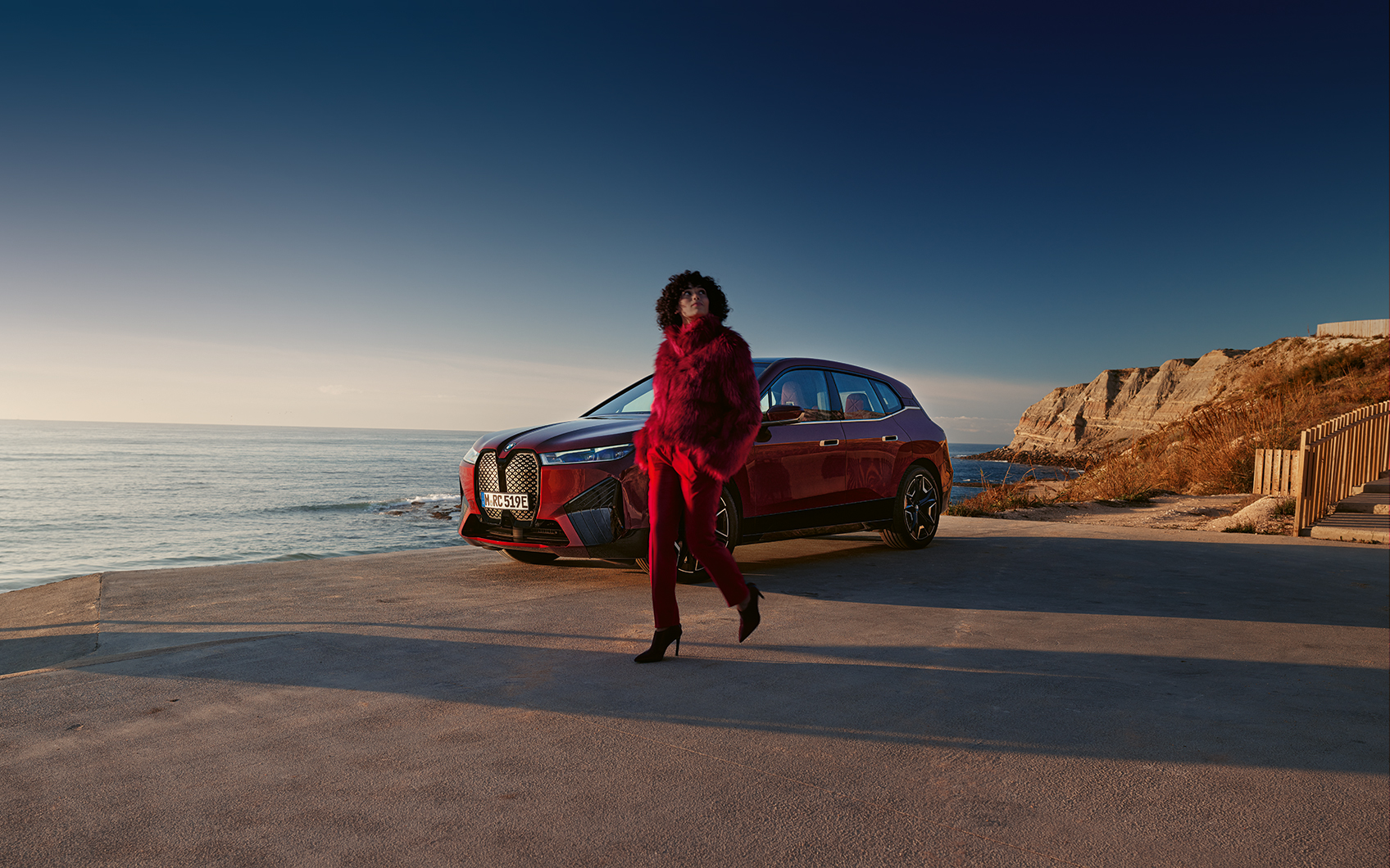 Rotes BMW iX Modell an Küstenstraße, Person in rotem Outfit, Meer und blauer Himmel