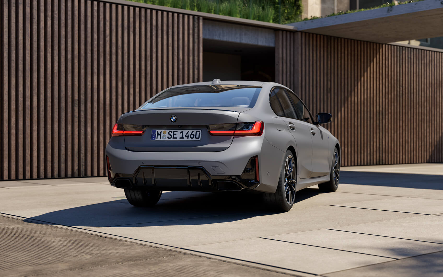Graue BMW M340i Limousine von hinten schräg auf Einfahrt vor moderner Holzfassade.