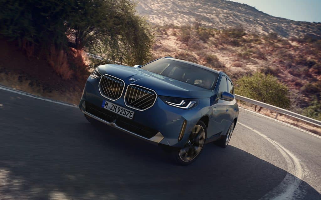 Frontansicht eines BMW X3 PHEV in der Farbe Blau beim bergauf Fahren auf einer Landstraße und Felsen im Hintergrund