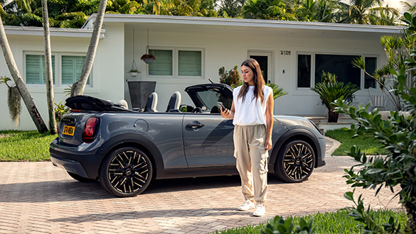 Graues MINI Cooper Cabrio mit geöffnetem Softtop-Verdeck in einer Einfahrt vor einem Wohnhaus; davor steht eine Frau, die auf ihr Handy schaut