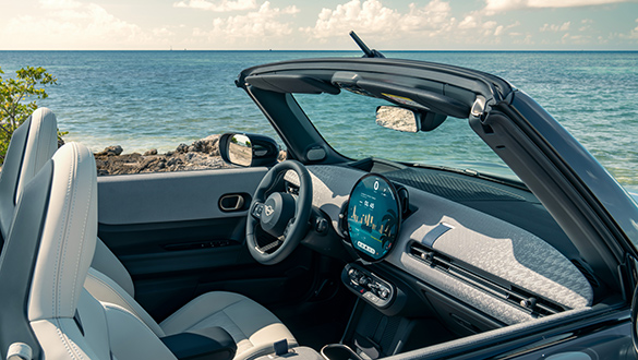 Interieur und Cockpit eines MINI Cooper Cabrio mit Sitzen aus beigem Vescin Kunstleder, Sport-Lenkrad und OLED Display vor einem Küstenhintergrund mit Blick auf das Meer