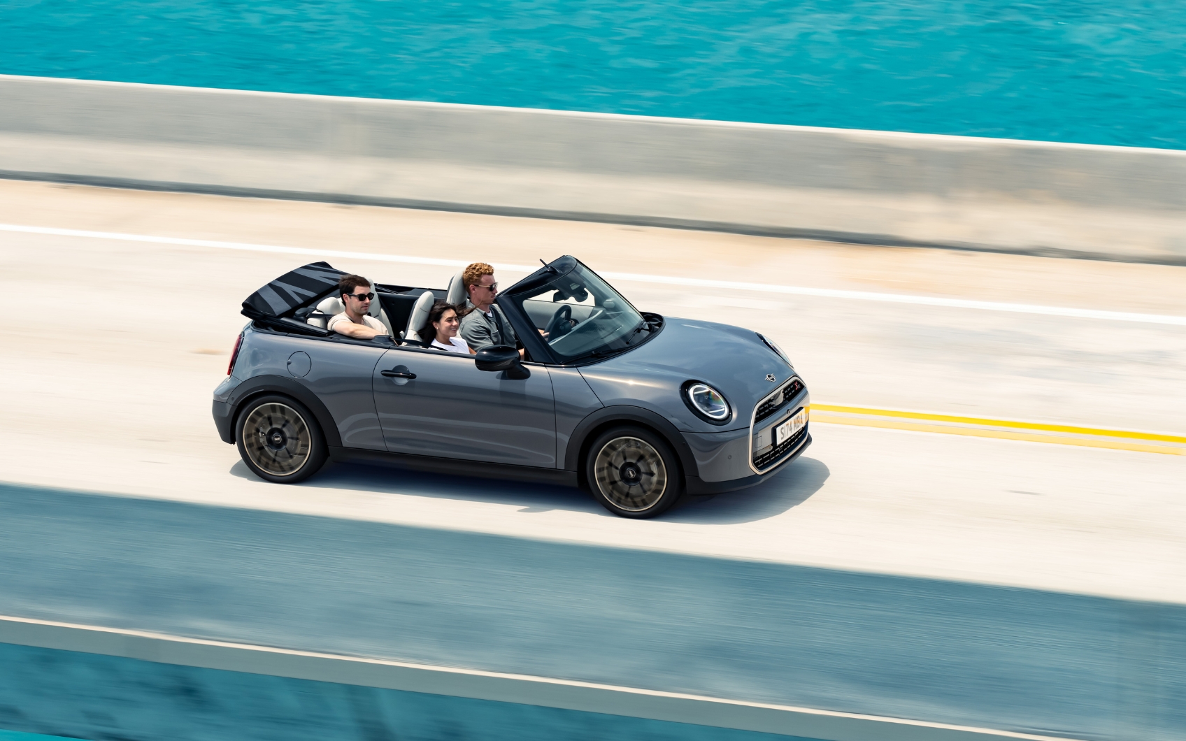 Blick von oben auf ein fahrendes MINI Cooper Cabrio in der Farbe Grau auf einer Küstenstraße mit Meer im Hintergrund