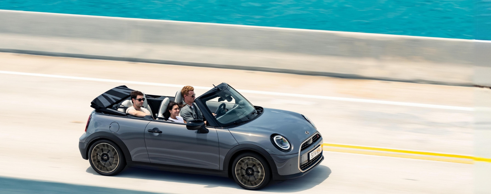 Blick von oben auf ein fahrendes MINI Cooper Cabrio in der Farbe Grau auf einer Küstenstraße mit Meer im Hintergrund