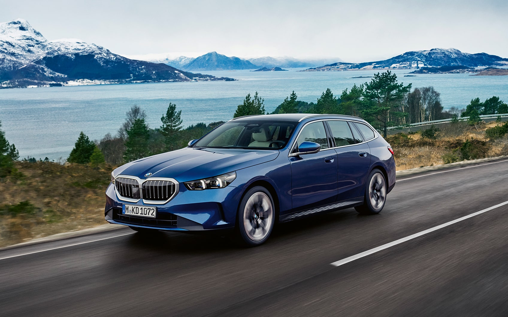 Blauer BMW 5er Touring fährt auf Landstraße mit See und verschneiten Bergen im Hintergrund.