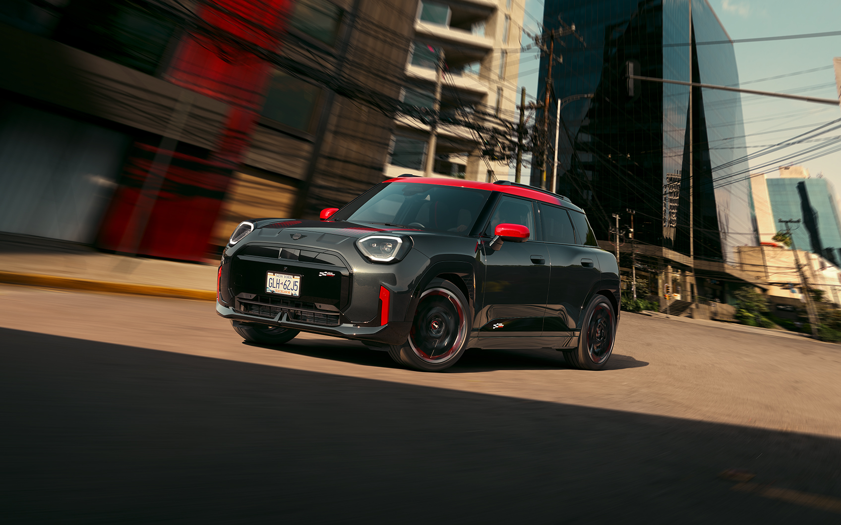 Profil und Frontansicht eines MINI John Cooper Works Aceman in der Farbe Schwarz mit roten Sport Stripes in Fahrt auf einer Straße in urbaner Umgebung