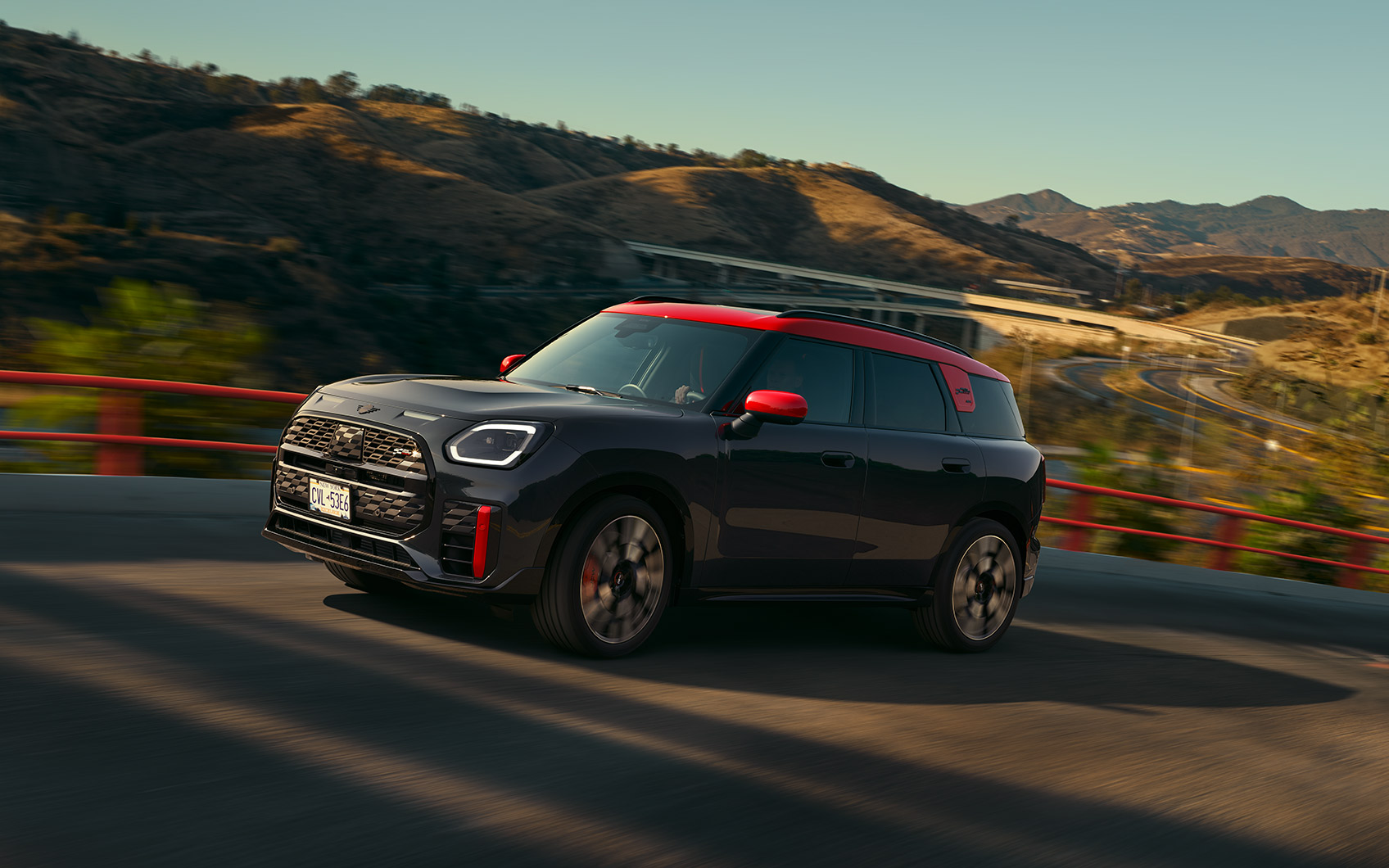Profil und Seitenansicht eines MINI JCW Countryman ALL4 in der Farbe Schwarz mit roten Sport Stripes und rotem Dach, fahrend auf einer Straße eines Berges