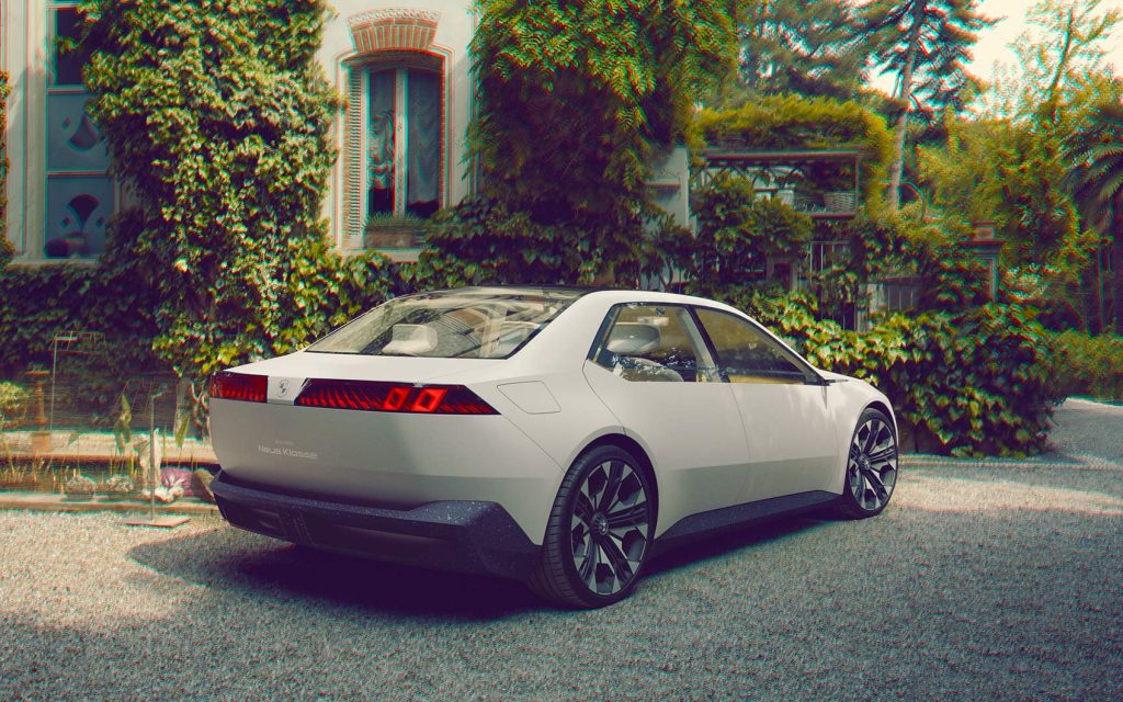 Weißes BMW Elektroauto mit Glasdach von schräg hinten, geparkt vor Hausfassade mit dichter Begrünung.
