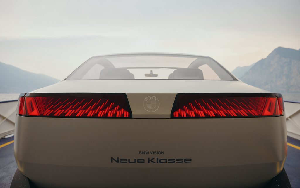 Rückansicht eines BMW Elektroautos mit leuchtenden LED-Rückleuchten, Schriftzug „Neue Klasse“ und Berglandschaft im Hintergrund.