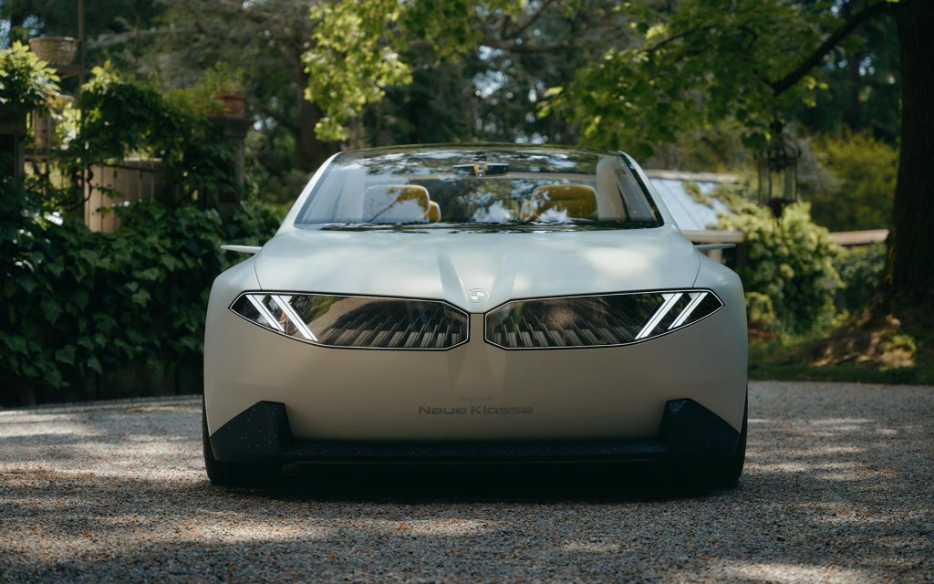 Frontansicht eines weißen Elektroautos mit auffälligem LED-Design, umgeben von Bäumen und Pflanzen.