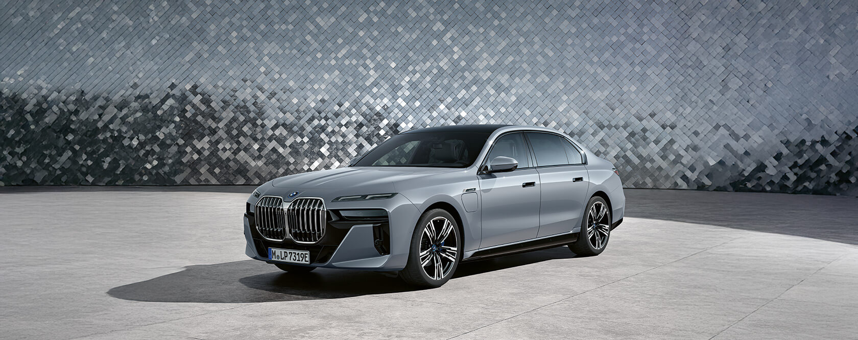 Silberne BMW 7er Limousine Plug-in-Hybrid mit geschlossenen Türen und markanter Front vor einer Wand mit silbernen Mosaikfliesen.