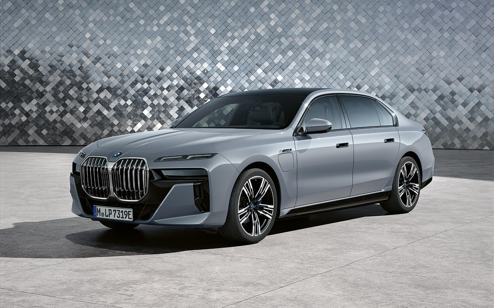 Silberne BMW 7er Limousine Plug-in-Hybrid mit geschlossenen Türen und markanter Front vor einer Wand mit silbernen Mosaikfliesen.