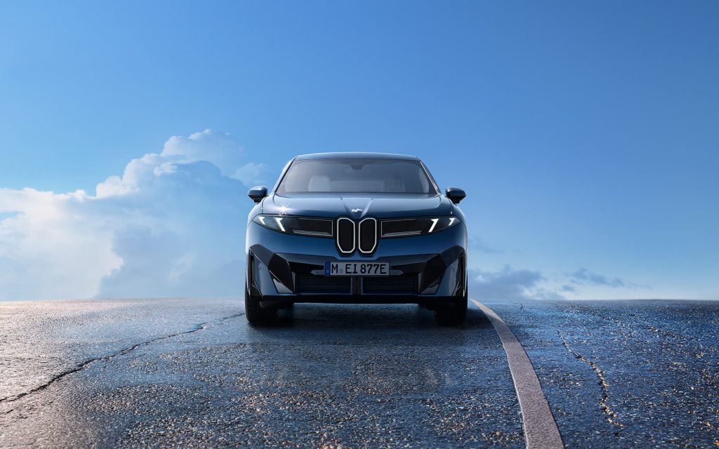 Blauer BMW iX3 steht frontal auf einer Straße. Im Hintergrund ist eine freundlicher blauer Himmel mit Wolken zu sehen.
