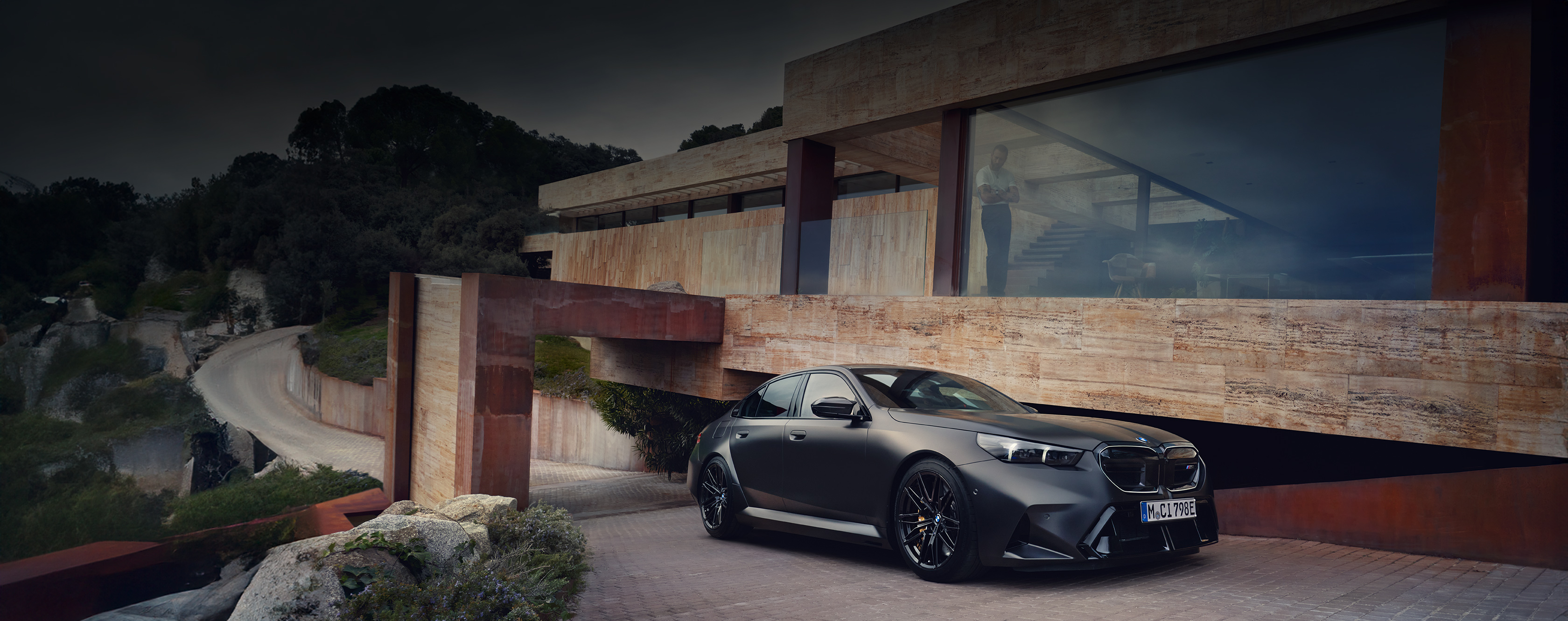 Nahaufnahme einer BMW M5 Limousine im Profil mit Blick auf die Front, Zufahrt eines modernen Wohnhauses mit Blick auf eine großen Glasfenster im Hintergrund der Limousine