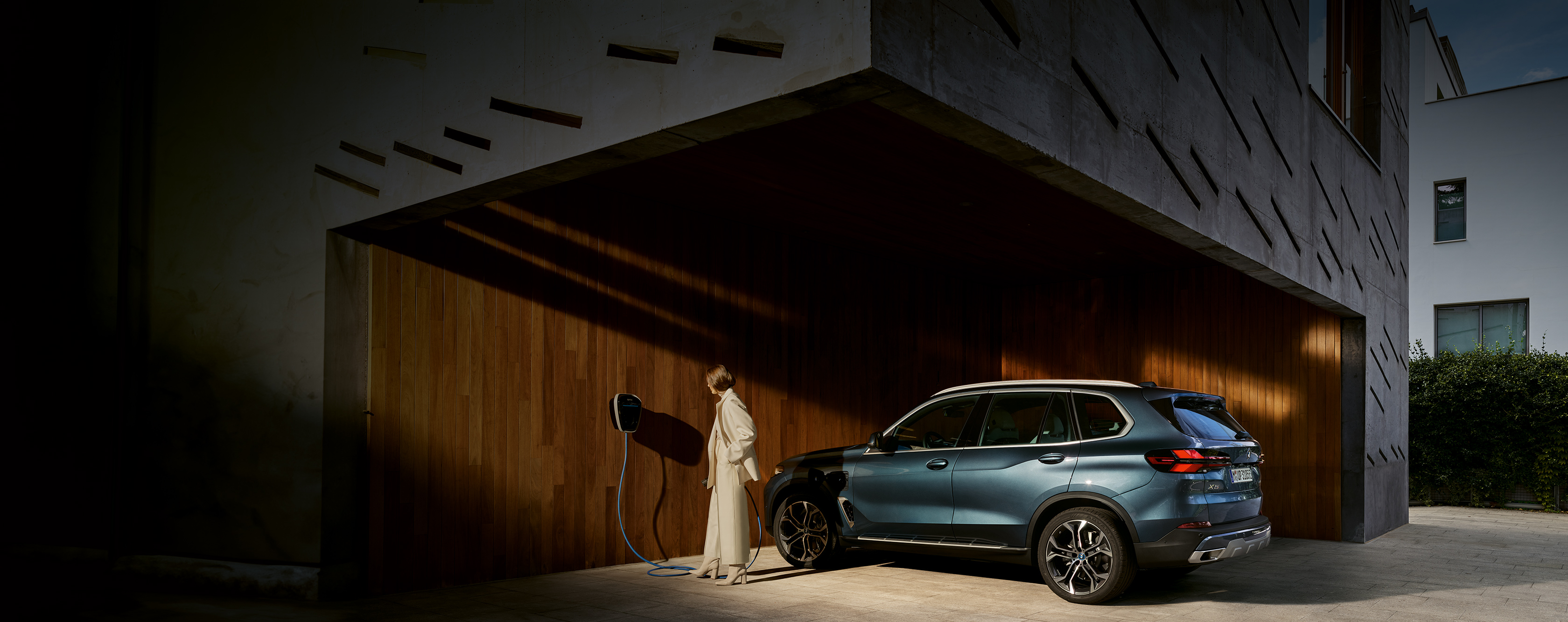 BMW X5 Plug-in-Hybrid steht an Gebäude. Frau im cremefarbenen Outfit will ihn über ein Kabel an die Ladebuchse anschließen