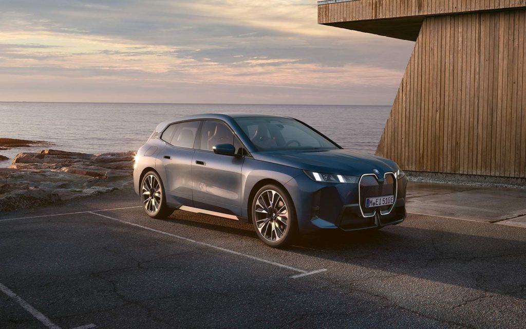BMW iX in der Farbe Blau auf einem Parkplatz geparkt mit Meer im Hintergrund, Fokus auf die Beifahrerseite des Fahrzeugs