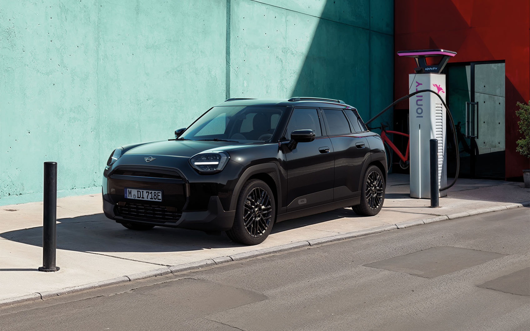 MINI Aceman Blackyard an Ionity-Ladesäule angeschlossen vor türkis-roter Hauswand.