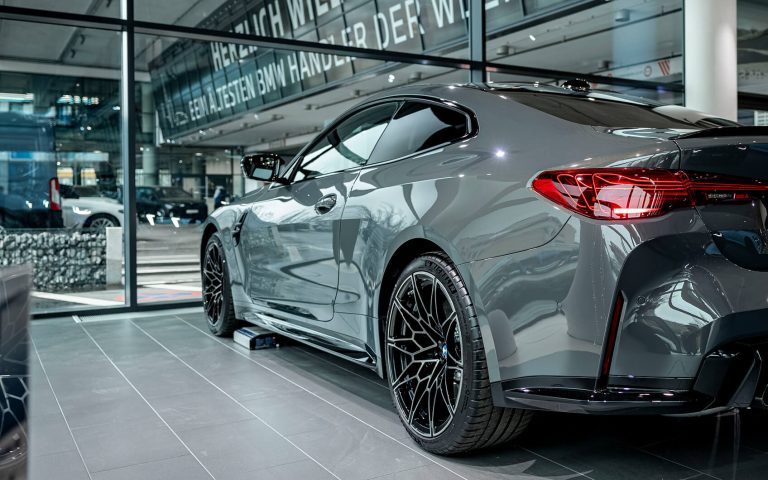 Graues BMW M4 Competition Coupé im Autohaus, Heckansicht schräg von links
