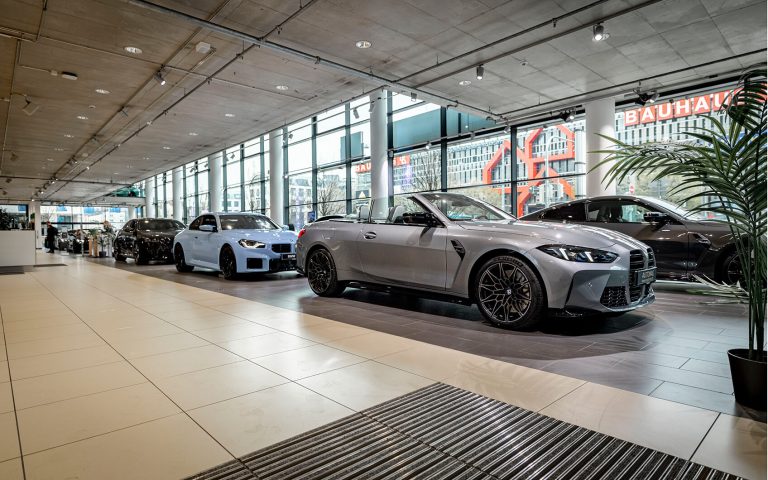 Graues BMW M4 Competition Cabrio im Autohaus, Frontansicht mit weiteren BMW Modellen
