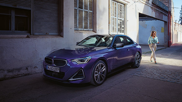 Lila BMW 2er Coupé parkt in urbaner Umgebung bei Sonnenschein, am Heck geht eine Frau vorbei