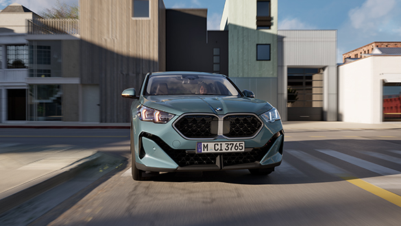 Grüner BMW X2 fährt dynamisch durch Stadtlandschaft, moderne Wohnhäuser im Hintergund
