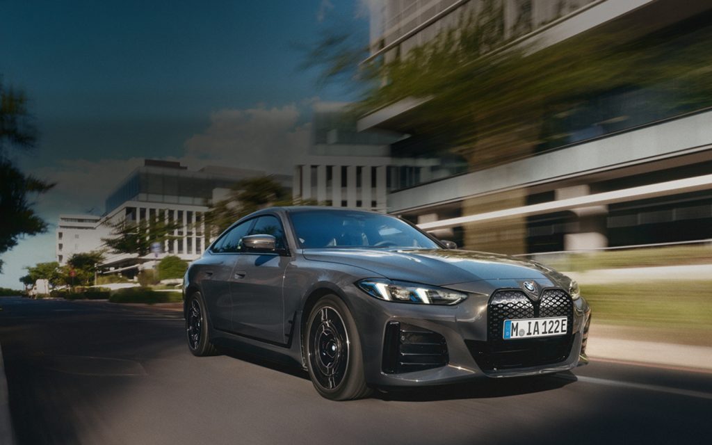 Grauer BMW i4 fährt dynamisch durch eine urbane Straße mit moderner Architektur im Hintergrund.