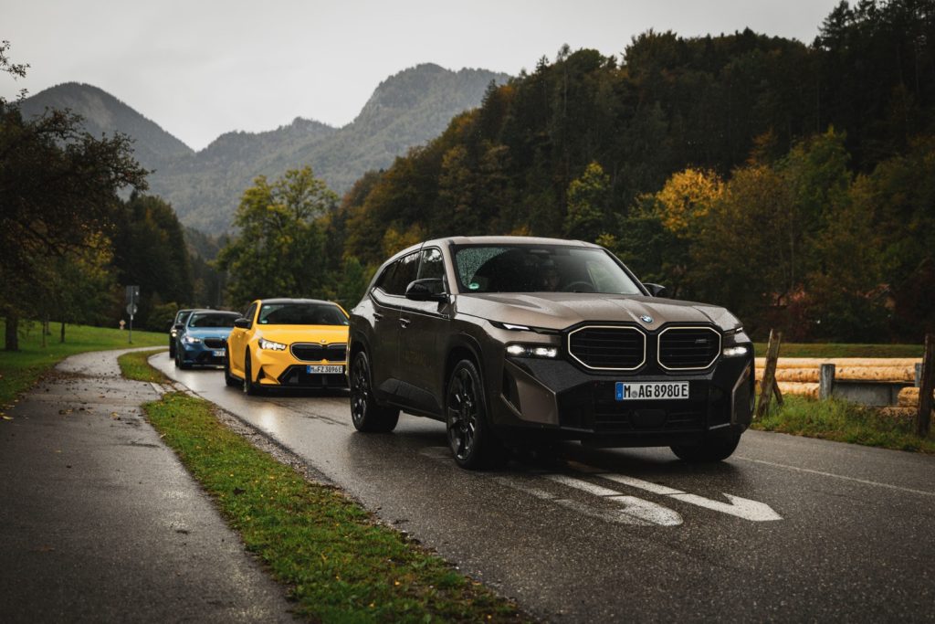 BMW XM SUV in Frontansicht, gefolgt von gelbem BMW M5 und blauem BMW M5 auf regennasser Landstraße.