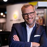 Christian Brandl lehnt mit verschränkten Armen in blauem Anzug am Bildschirmrand