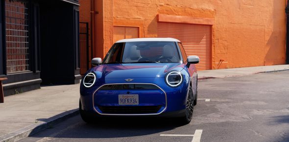 Blauer Mini Cooper Electric mit weißem Dach frontal geparkt vor orangefarbener Wand.
